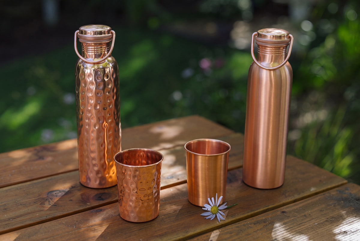 Garrafas de cobre, copos lisos e martelados e uma margarida numa mesa de jardim de madeira