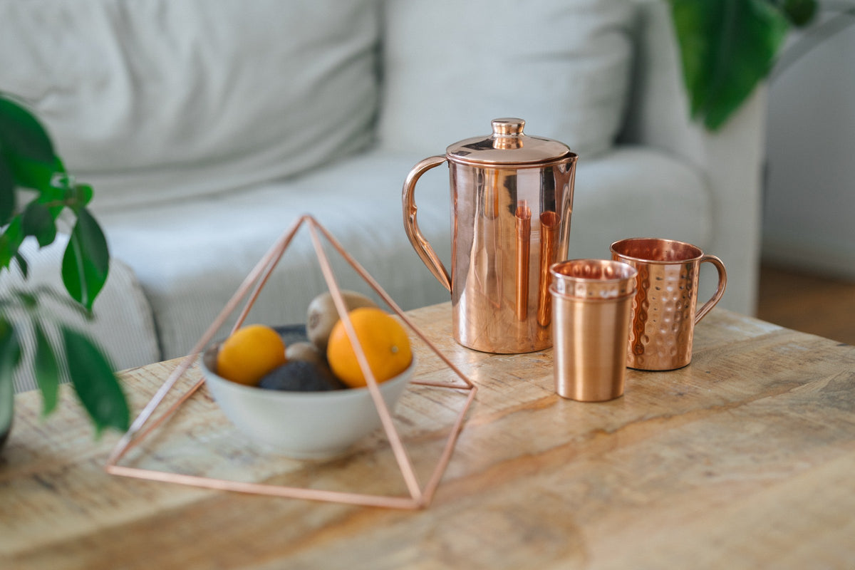 Pirâmide de cobre sobre uma taça de fruta, com um jarro, um copo e uma caneca numa mesa de centro de madeira