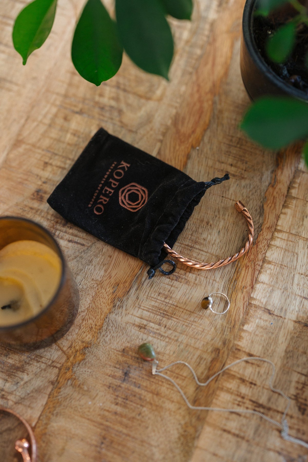 Kopero Pulseira de cobre Trançado, sobre uma mesa de madeira ao lado do saco de veludo, de uma vela e de joias