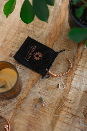 Kopero Pulseira de cobre Trançado, sobre uma mesa de madeira ao lado do saco de veludo, de uma vela e de joias