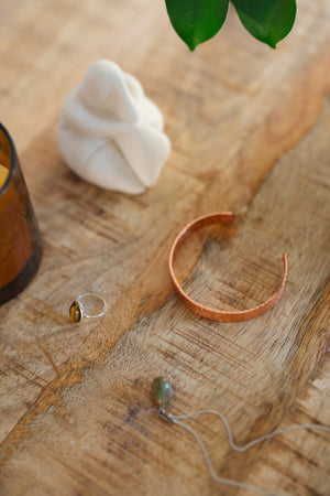 Kopero Pulseira de cobre Martelado, sobre uma mesa de madeira ao lado de uma figura e de um anel