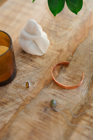Kopero Pulseira de cobre Liso, sobre uma mesa de madeira ao lado de uma figura e de um anel