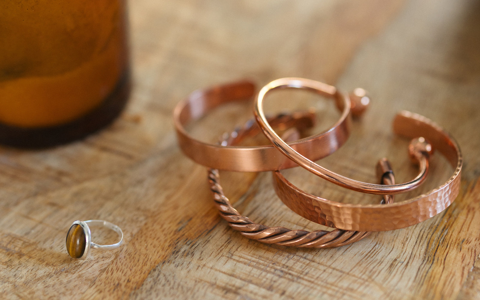Quatro pulseiras de cobre, trançada, lisa, martelada e redonda, numa mesa de madeira ao lado de uma vela e de um anel