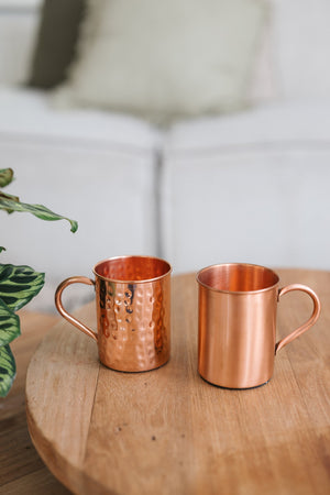 Kopero Caneca de cobre, uma caneca lisa e uma martelada lado a lado sobre uma mesa redonda de madeira