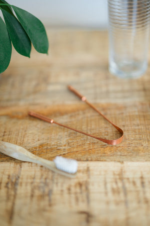 Kopero Raspador de língua de cobre, sobre uma mesa de madeira ao lado de uma escova de dentes de bambu e de um copo de água