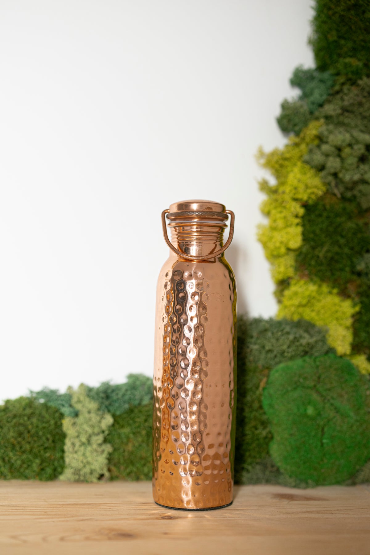 Garrafa de água de cobre Kopero Martelada, 900 ml, na vertical sobre uma mesa de madeira com um fundo de musgo