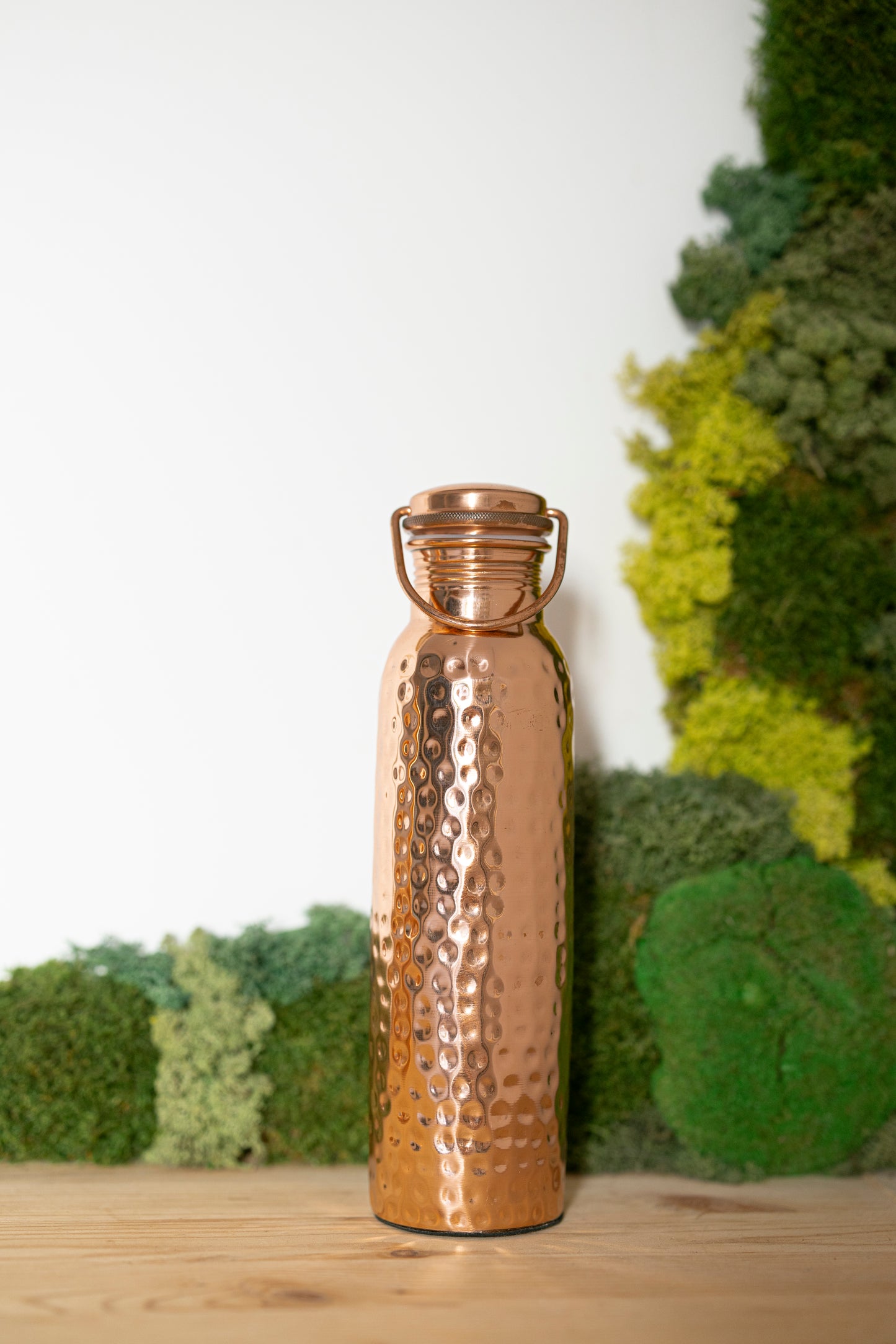 Garrafa de água de cobre Kopero Martelada, 900 ml, na vertical sobre uma mesa de madeira com um fundo de musgo