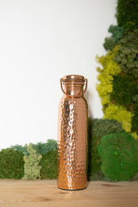 Garrafa de água de cobre Kopero Martelada, 900 ml, na vertical sobre uma mesa de madeira com um fundo de musgo
